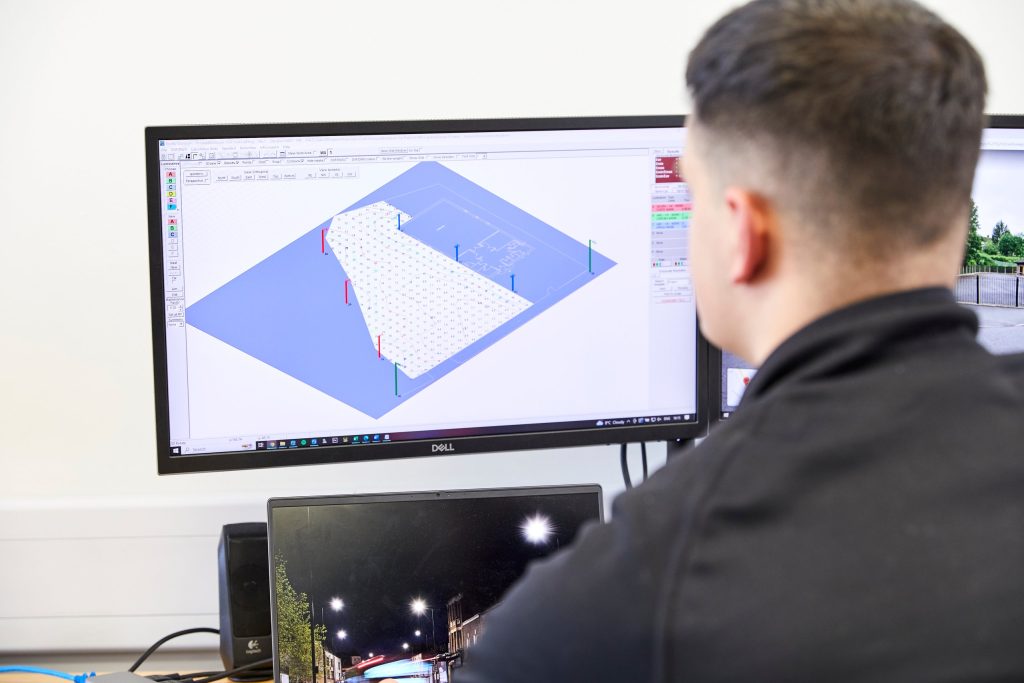 An INDO lighting designer working on a street lighting calculation, with a night street photograph on the second screen